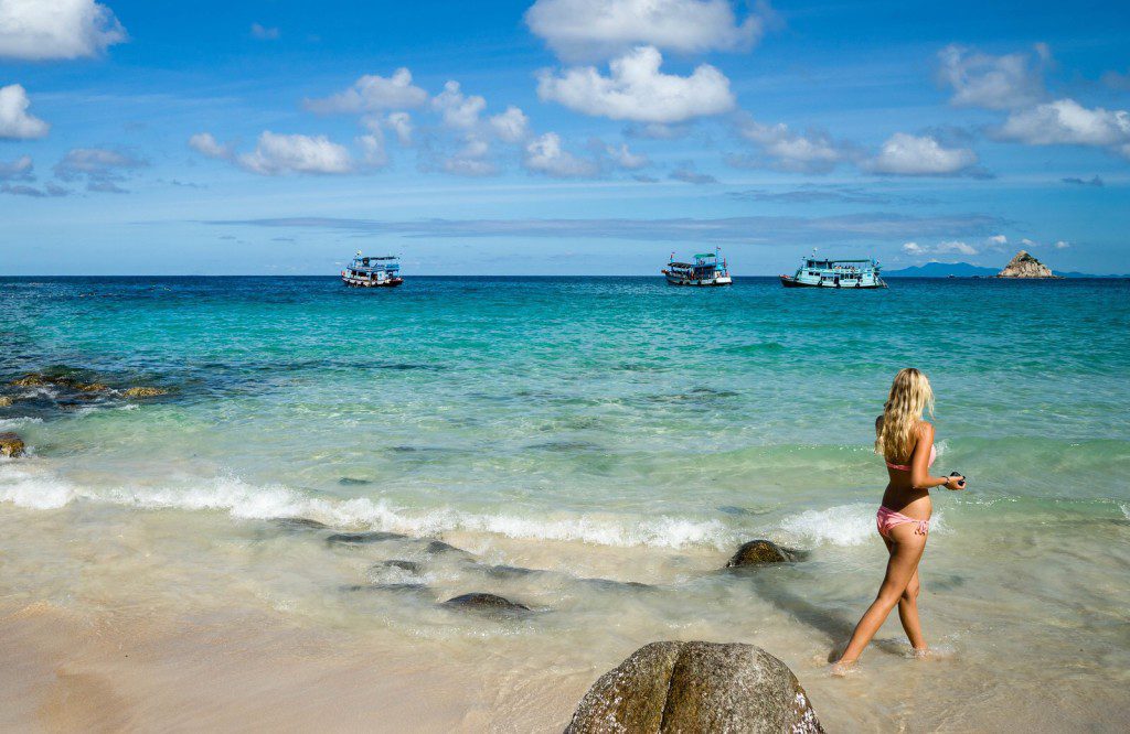 Koh Tao's Best Beaches – Tanote Beach. A person stands on a sandy beach looking at the clear turquoise sea with boats floating and a small island in the distance under a blue sky.