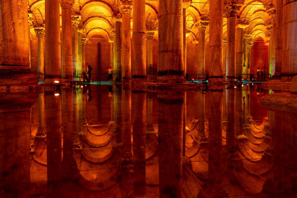 This image shows the interior of an ancient cistern with illuminated columns reflecting on the calm water surface, creating a symmetrical, serene, almost mystical ambiance.