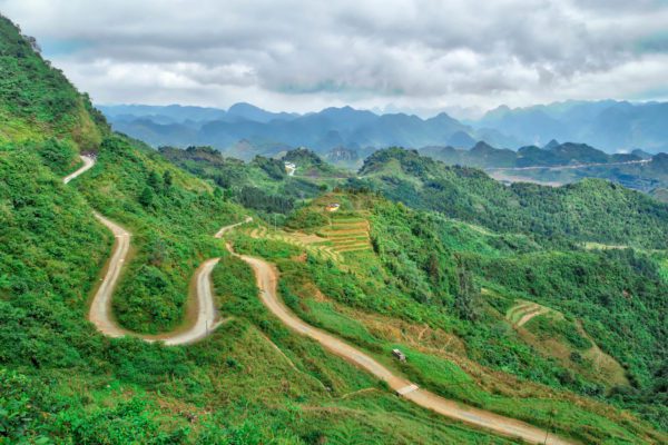 A winding road snakes through a lush green mountainous landscape with terraced fields. Overcast skies loom above this serene, hilly terrain.