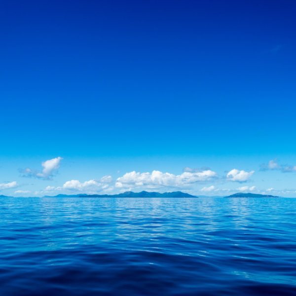 This image depicts a calm blue ocean under a clear sky, with scattered clouds above. A faint silhouette of an island is visible on the horizon.