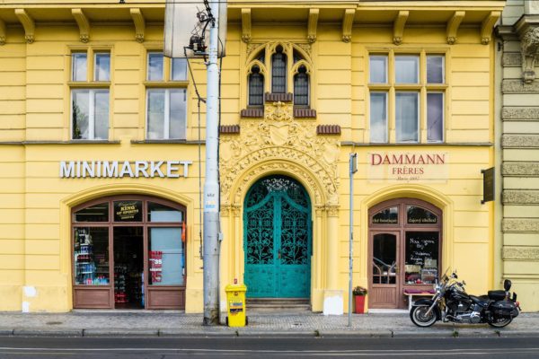 A yellow European-style building with two shop fronts, featuring a Minimarket and Dammann Frères. A motorcycle is parked outside beside a mailbox.