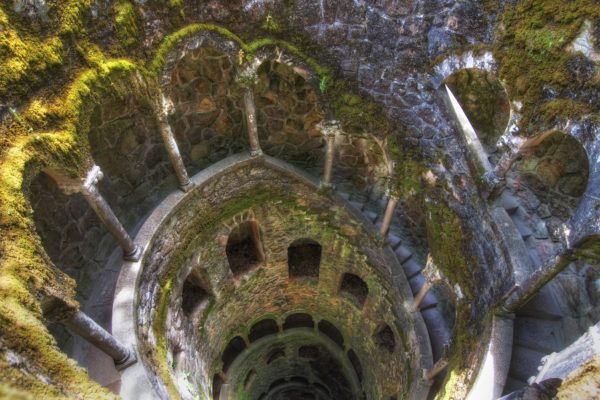 An ancient stone well with moss overgrowth, viewed from above, showing circular stairs descending around the periphery into the shadowy depths below.