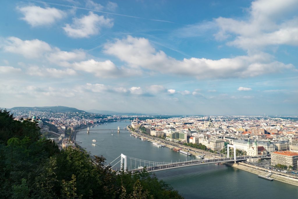 A panoramic view of a cityscape with a river flowing through, multiple bridges crossing over, buildings lining the banks, and hills in the background.