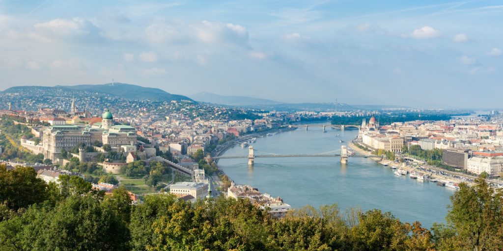 This is a panoramic view of a city with a river bending through it, crossed by bridges, framed by green hills and a mix of historic and modern buildings.