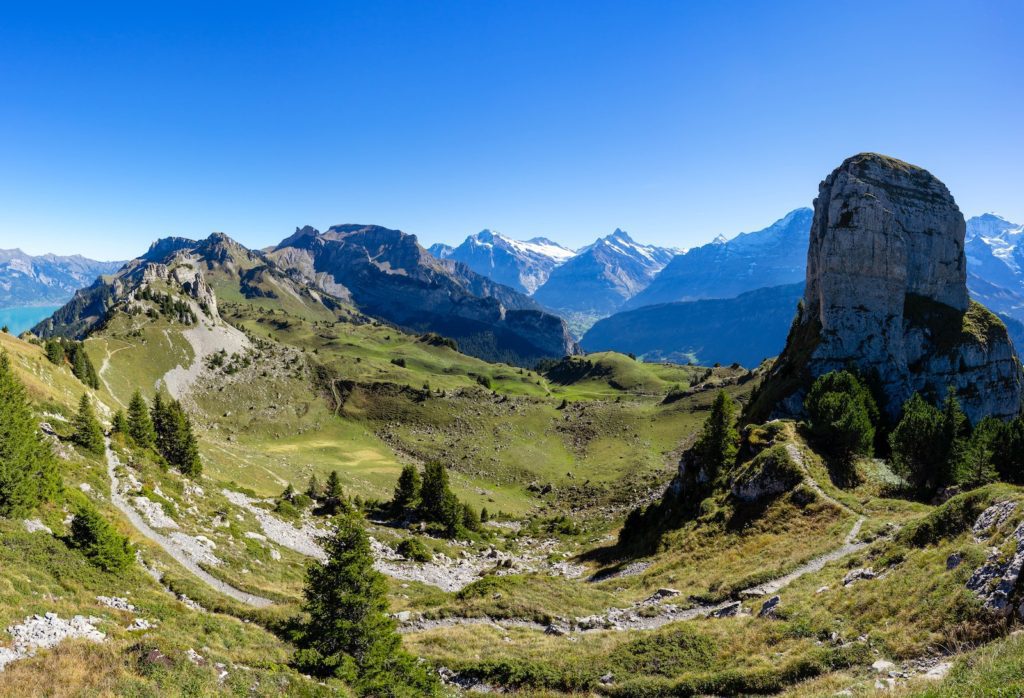 A panoramic view of a mountainous landscape featuring a towering rock formation, rolling green hills, hiking paths, and distant snow-capped peaks under a clear blue sky.