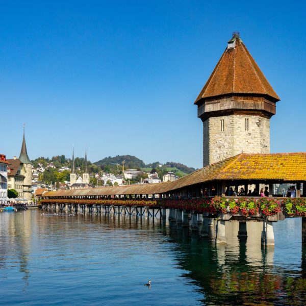 This image shows the Chapel Bridge and Water Tower in Lucerne, Switzerland, reflecting in clear water with colorful flowers and traditional architecture.