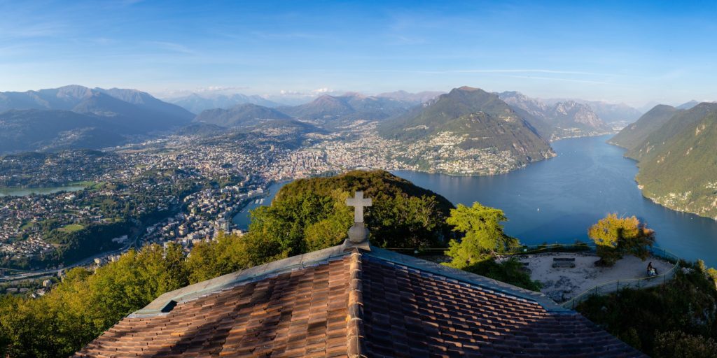 This panoramic image features a tranquil lake surrounded by mountains, with a town spread along its shore, viewed from a high vantage point with a cross.