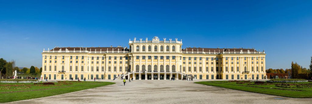 This image shows a grand, historic palace with ornate architecture, large windows, and statues atop its roof, under a clear blue sky, surrounded by manicured lawns.