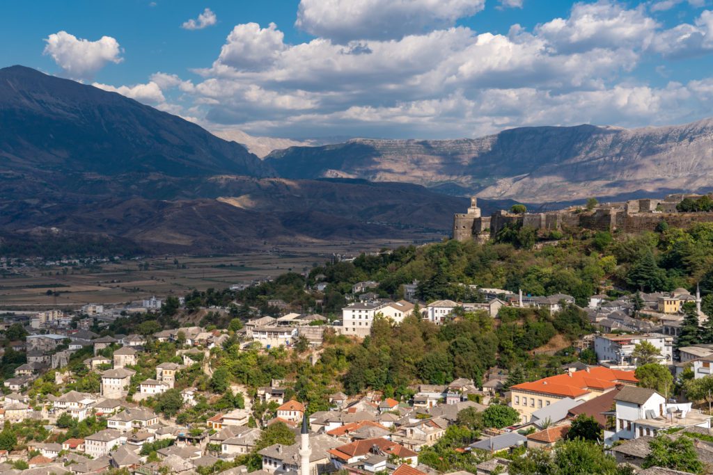 A scenic view of a historic town with dense buildings, a prominent castle, surrounded by mountains under a partly cloudy sky.