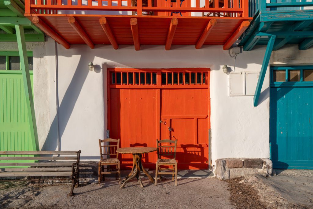 A colorful, quaint scene with a vibrant red door, matching balcony, green shutters, and blue accents. Wooden chairs invite relaxation under the Mediterranean sun.