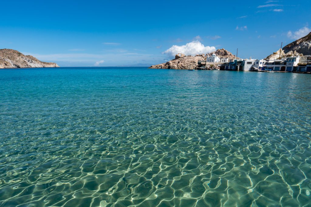 Crystal-clear blue water leads to a coast with buildings and a dock. Rugged hills under a blue sky with wispy clouds complete the tranquil scene.