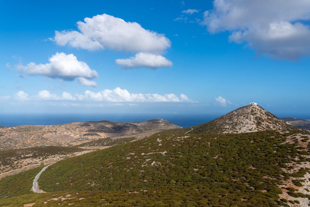 A scenic landscape featuring a winding road leading up to a hilltop. Lush greenery blankets the slopes, with the sea and blue skies in the background.