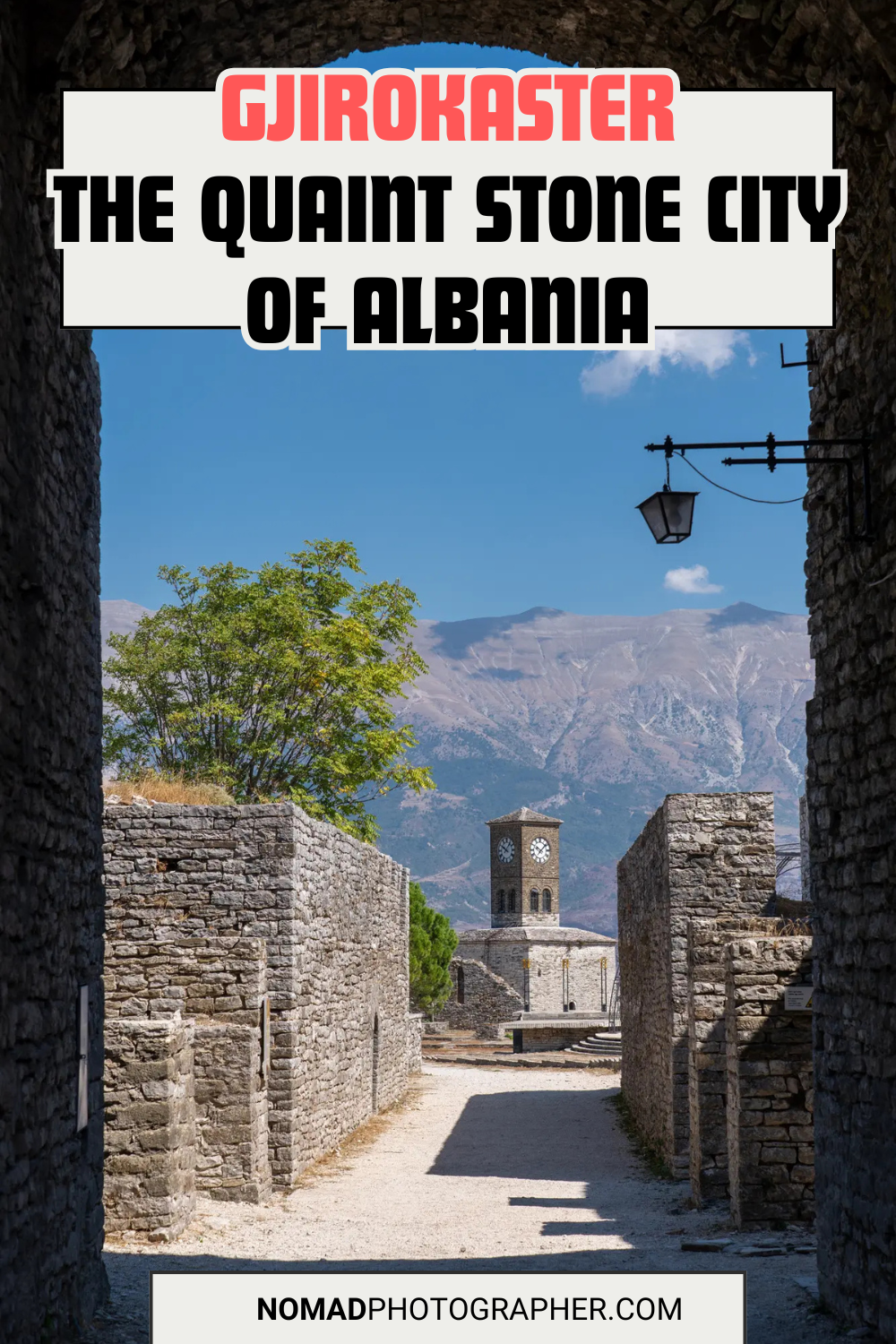 Historic stone buildings in Gjirokastër, Albania, with a clock tower in the background and mountainous landscape. A path leads through weathered stone walls.