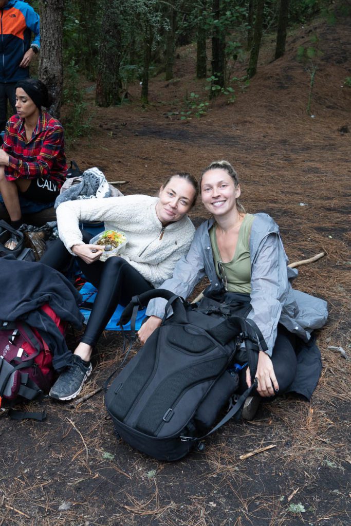 Two people are smiling and sitting on the ground in a forest with backpacks. One person is eating. Another person is in the background.