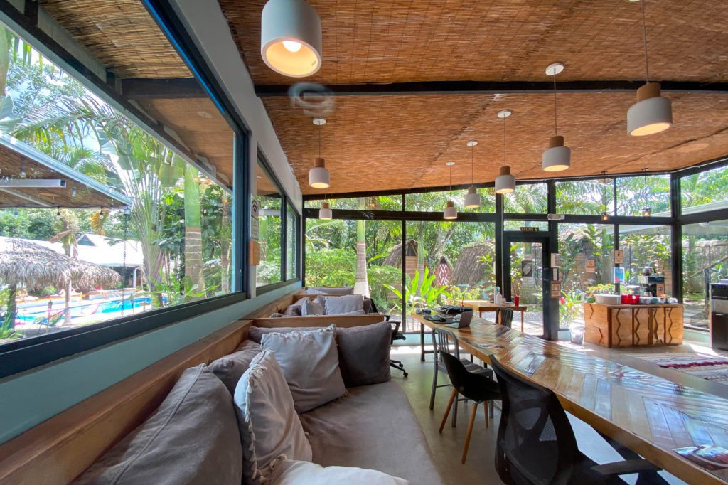 This is a modern, tropical coworking space for digital nomads. With a long wooden table, comfortable seating, and pendant lights, surrounded by large windows reveal lush greenery outside.