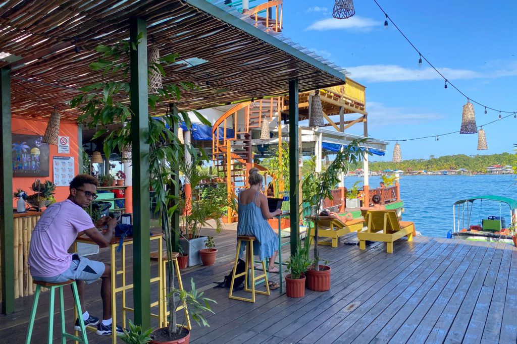 A vibrant waterfront dining area with people at tables, a wooden deck, green potted plants, string lights, and a view of blue water.