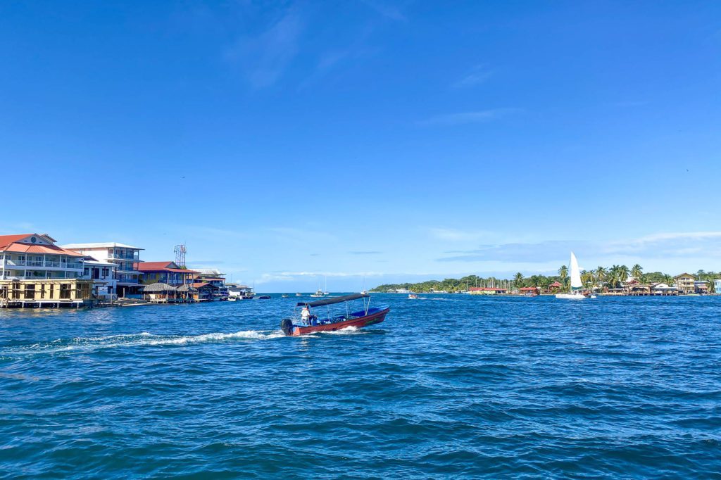 A small boat with two persons speeds across a clear blue sea with waterfront buildings under a sunny sky, possibly in a tropical locale.