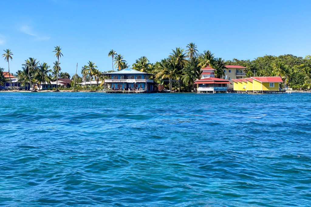 A tropical scenery with clear blue water and colorful buildings along the shoreline. Palm trees are visible under a bright blue sky.