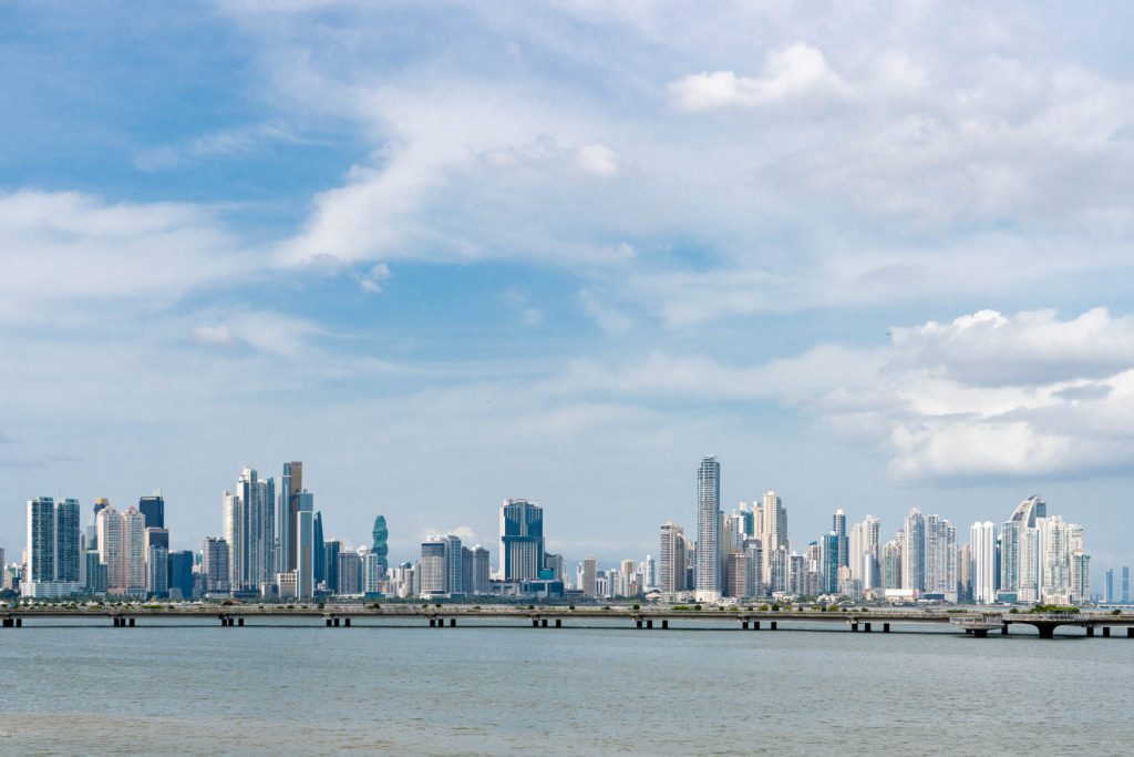 A panoramic daytime skyline of a modern city with numerous skyscrapers, viewed across a wide river with a bridge in the foreground under a partly cloudy sky.