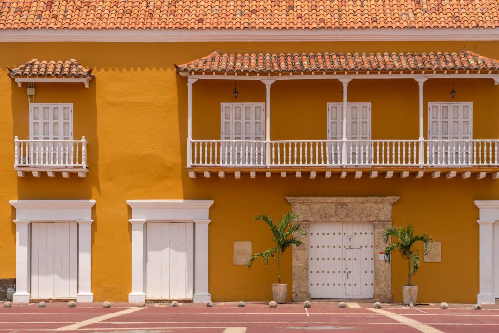 A bright yellow colonial building with a red-tiled roof features white framed doors and windows, two balconies, and decorative plants by the entrance.