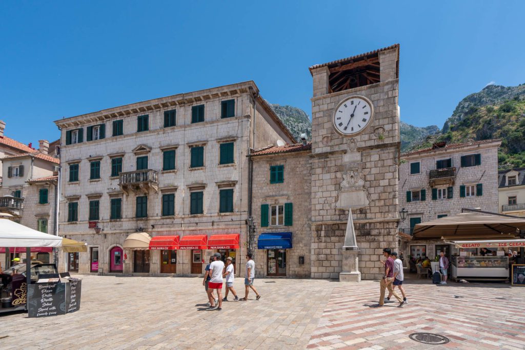 The image shows a sunny town square with historical buildings, a clock tower, and a few people walking. Mountains are visible in the background.