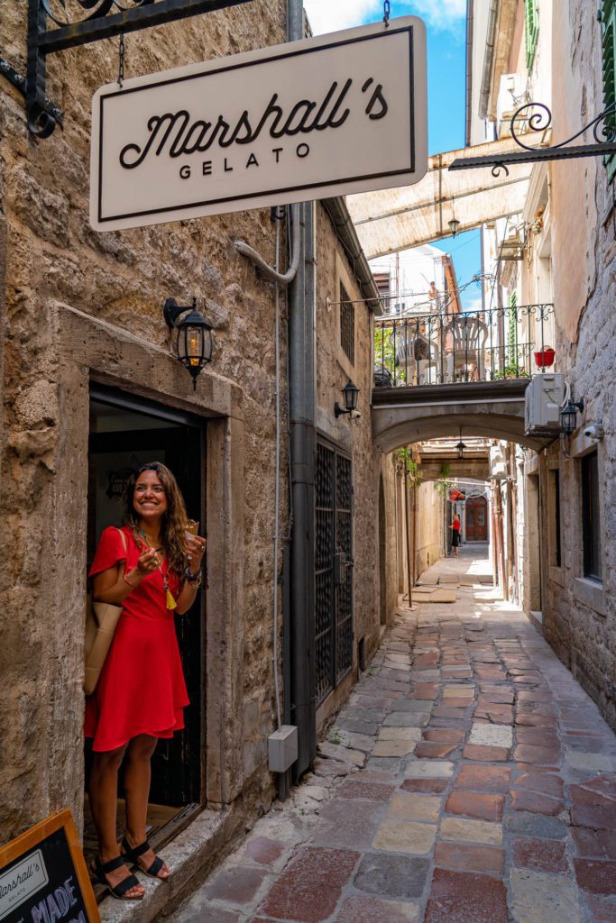 A person in a red dress stands smiling at a gelato shop doorway under a sign, in a narrow, picturesque cobblestone alley with old stone buildings.