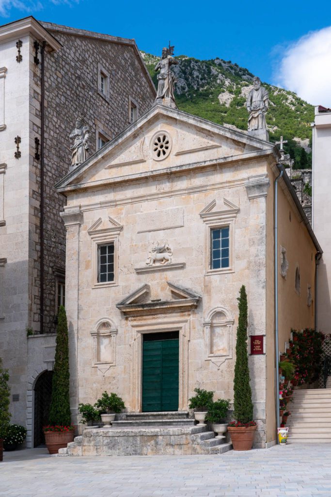 This is a historic stone church with a triangular pediment, green door, statues, and steps, nestled between buildings against a mountainous backdrop.