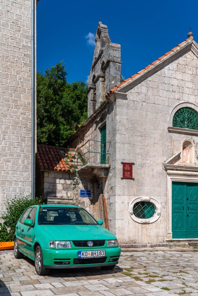 A green car is parked in front of a traditional stone building with a red-tiled overhang, round window, and a bright green door under a clear blue sky.