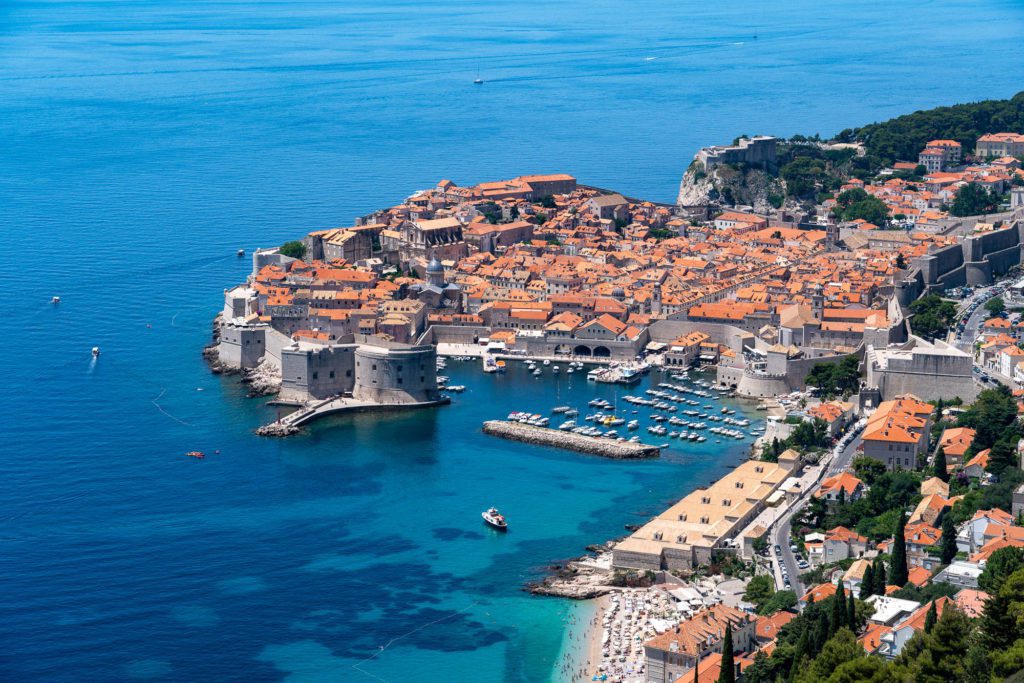 An aerial view of a coastal old city with terracotta rooftops, ancient walls, a marina with boats, adjacent to a deep blue sea.