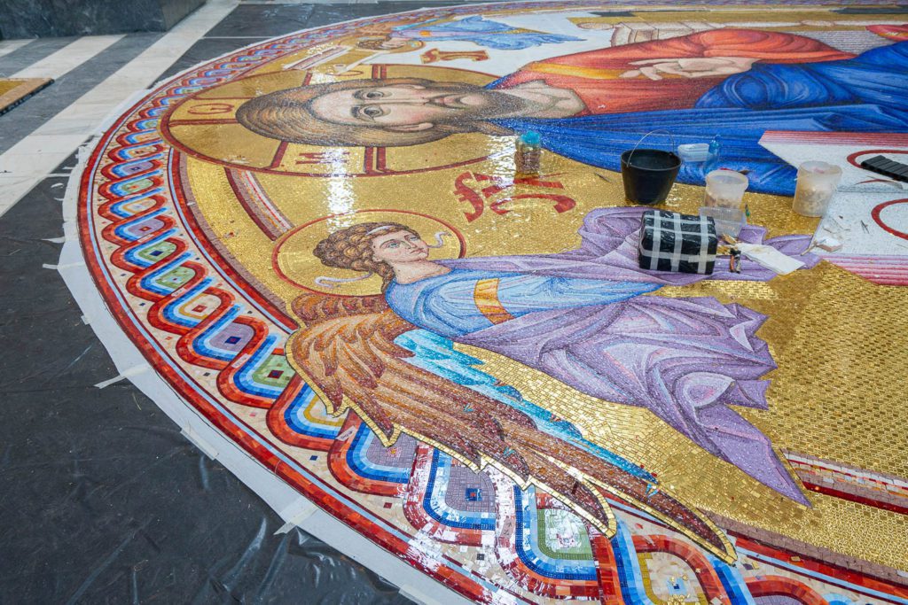 This image shows a large, intricately detailed mosaic artwork in progress, featuring religious figures with vibrant colors and golden backgrounds, along with artist materials.