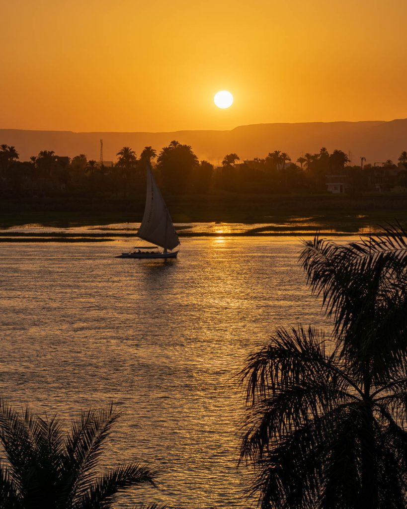 A sailboat glides on calm water at sunset, with the sun hovering low over distant hills. Palm tree silhouettes frame the tranquil, golden scene.