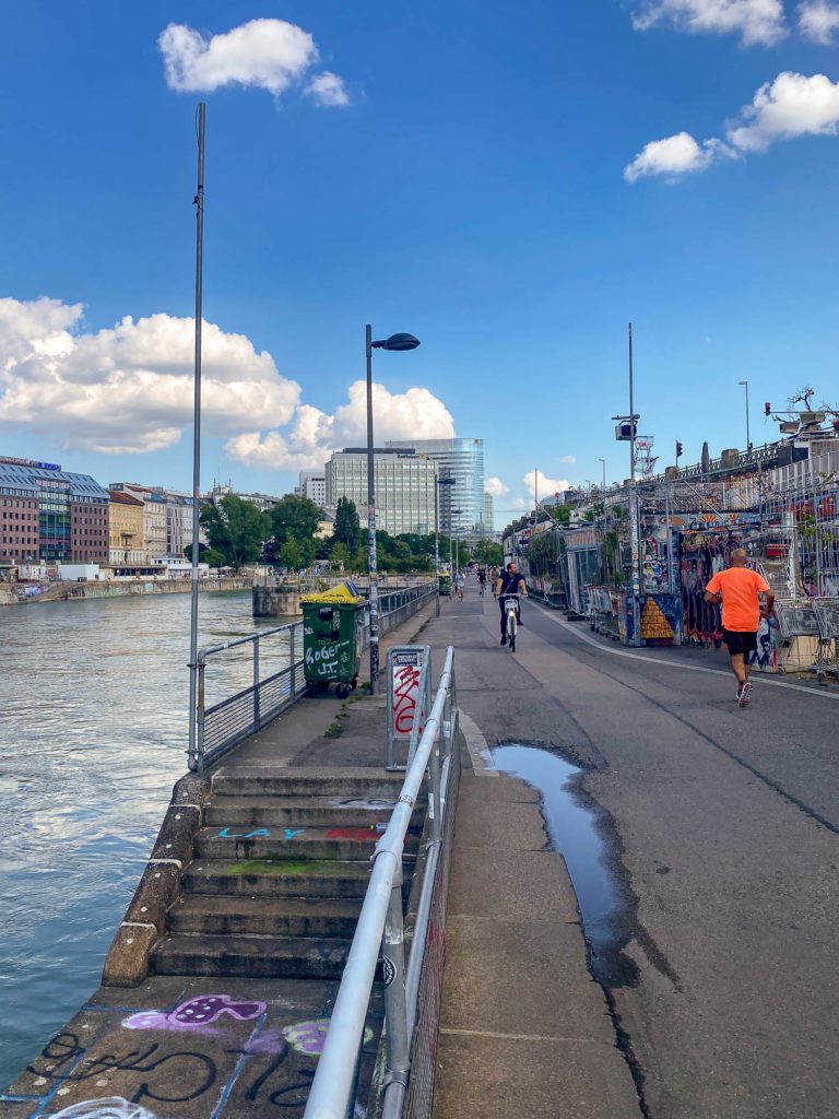 A riverside walkway with two people, one cycling and one walking in Vienna Austria. Graffiti, urban art, and market stalls line the path under a blue sky with clouds.