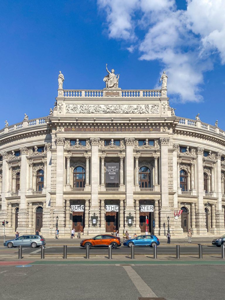 An ornate historical building with classical architecture under a blue sky with clouds in Vienna Austria. People and cars are visible in front, behind a metal barrier.