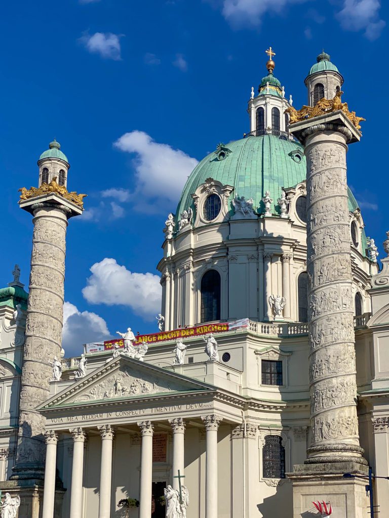 The image shows an ornate baroque-style church with two tall columns in the foreground against a bright blue sky with scattered clouds.