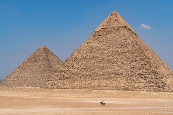 The image shows two ancient Egyptian pyramids under a clear blue sky, with a person riding a horse in the vast desert foreground.