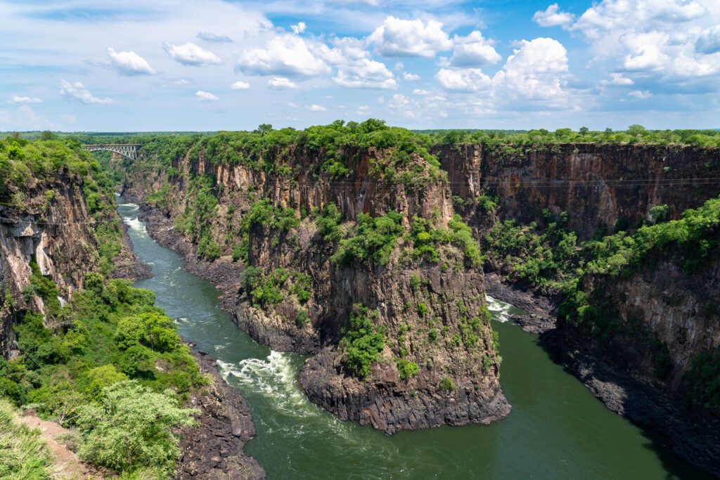 A majestic gorge with steep, rocky cliffs and lush vegetation overlooking a winding river under a blue sky with scattered clouds. A bridge spans the distance.