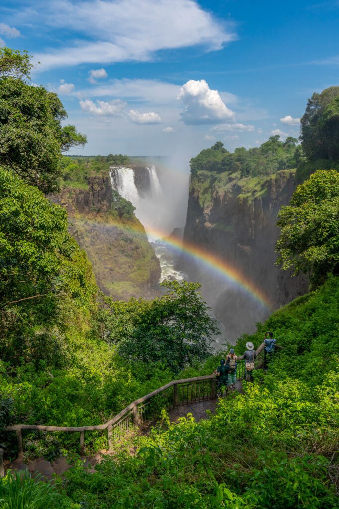 A majestic waterfall surrounded by lush greenery with a vibrant rainbow arcing across the mist. People observe the scenic beauty from a viewing platform.