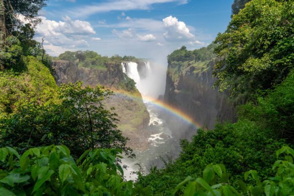 A majestic waterfall plunges between lush, green cliffs, creating a mist that gives rise to a vibrant rainbow under a sunny, partly cloudy sky.