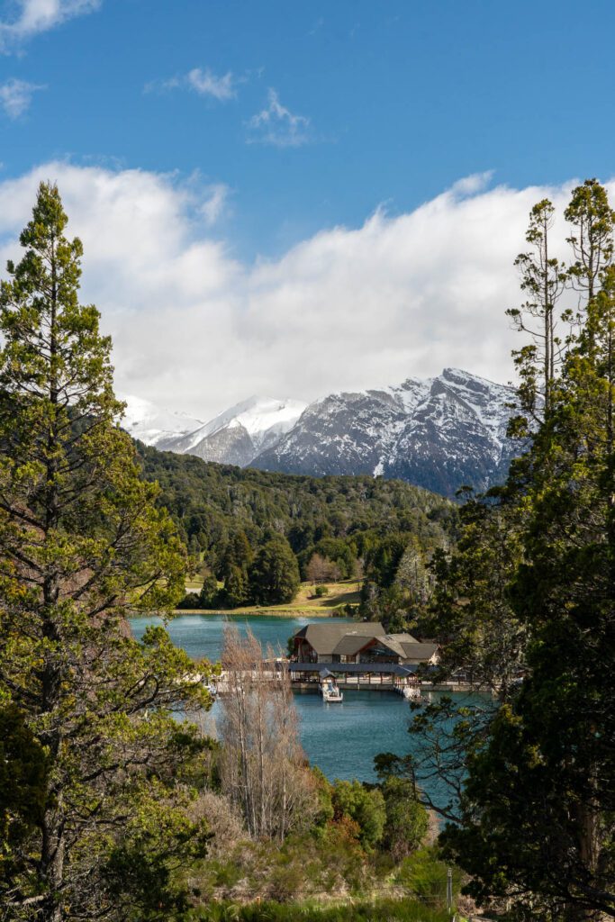 A tranquil lake with a wooden structure nestled amidst greenery, framed by trees, with a backdrop of majestic snow-capped mountains under a blue sky with clouds.