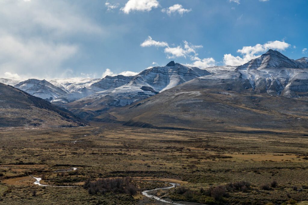 A winding road meanders through a vast valley leading towards snow-capped mountains under a partly cloudy sky, highlighting the rugged natural landscape.