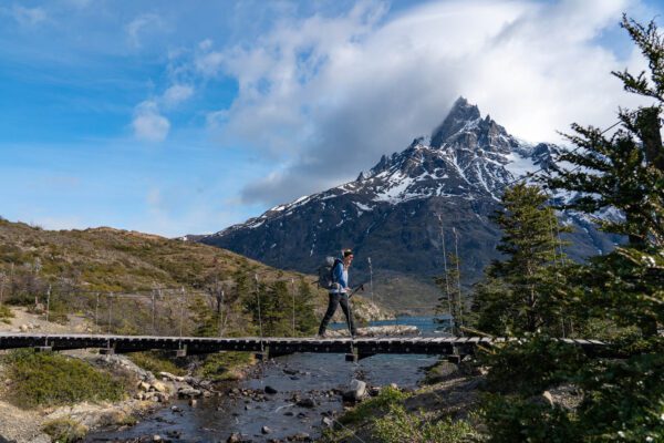 A person is hiking across a wooden boardwalk with scenic mountains in the background, under a blue sky with wispy clouds.
