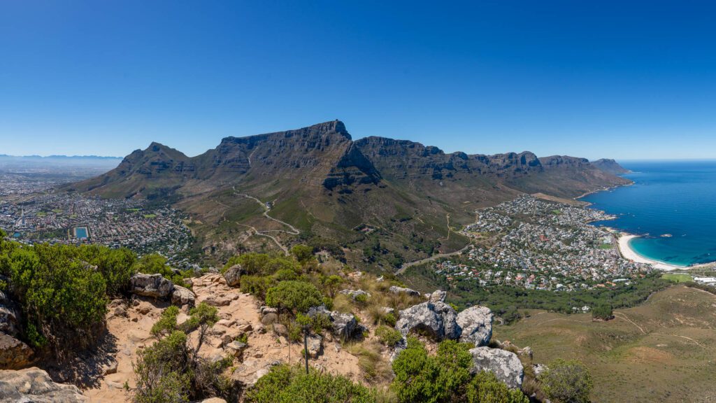 Panoramic view overlooking a coastal city with a prominent flat-topped mountain range, lush greenery, urban architecture, and a deep blue ocean horizon.