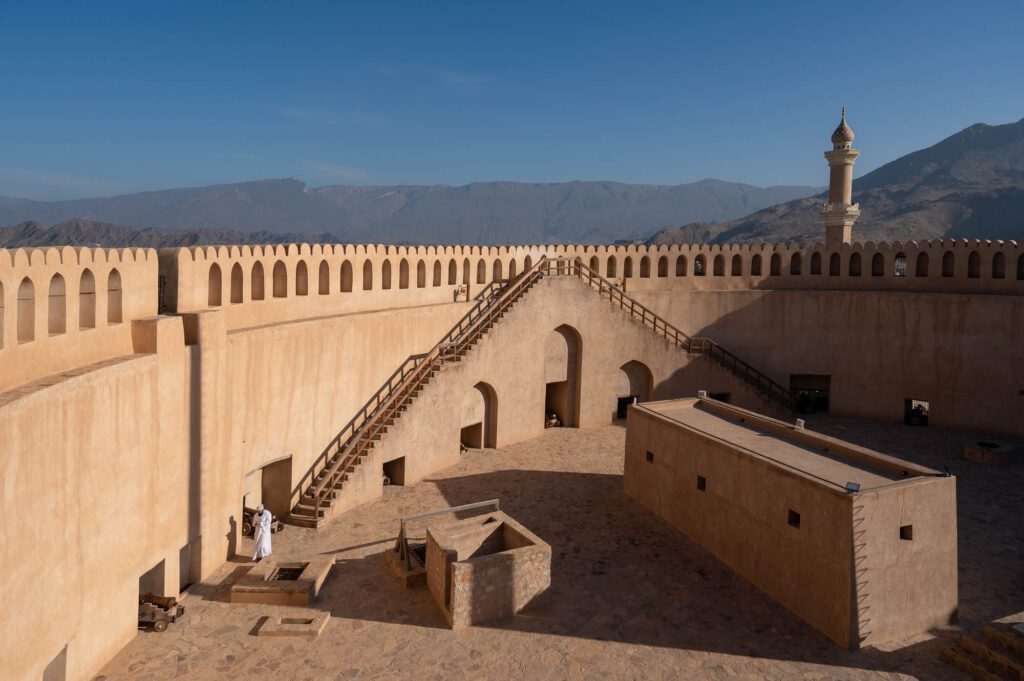 The image shows an ancient fort with crenellated walls, a minaret, and stairs leading upwards. It's a sunny day with clear blue skies and mountainous background.