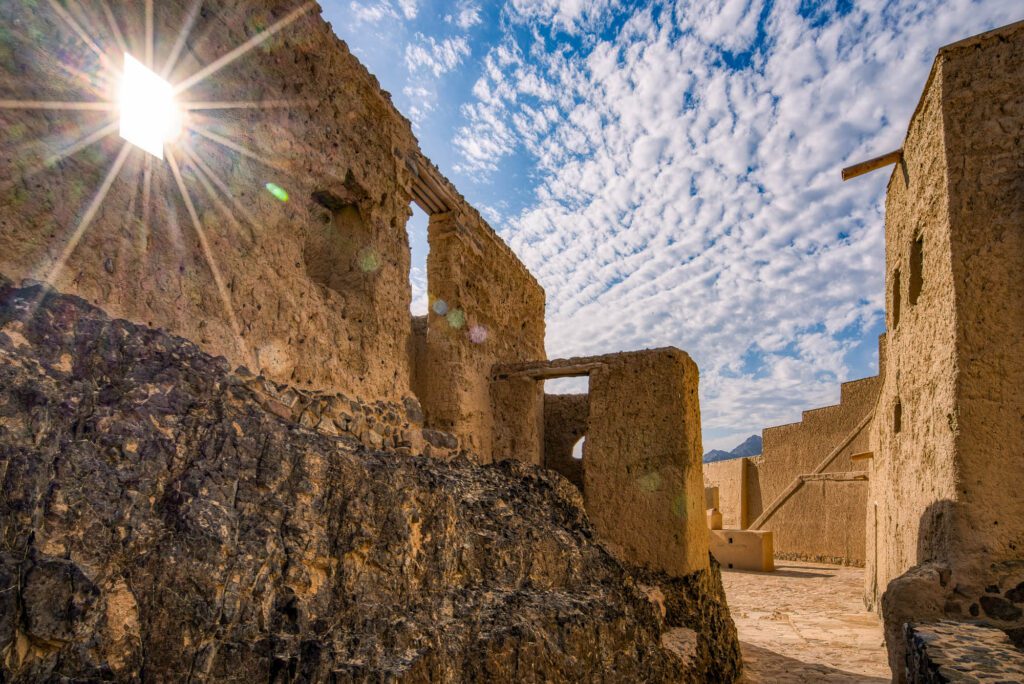 The image features ancient stone structures under a bright sun with a backdrop of blue sky and wispy clouds. Weathered walls suggest historical significance.