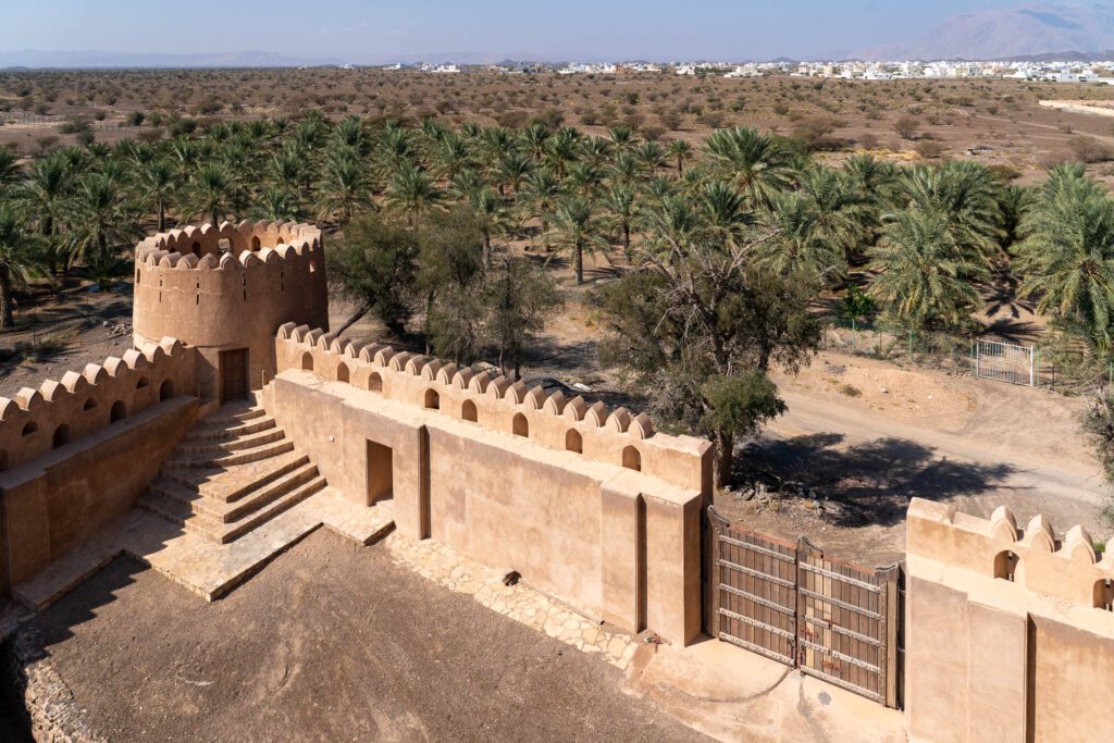 This is an aerial view of a desert fortification with crenellated walls, overlooking a lush palm grove. A settlement is visible in the distance under a blue sky.