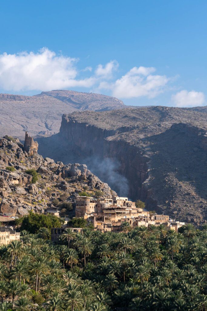 A village nestled on a mountain slope above a dense palm grove, with smoke rising from a structure against a backdrop of rugged cliffs and a blue sky.