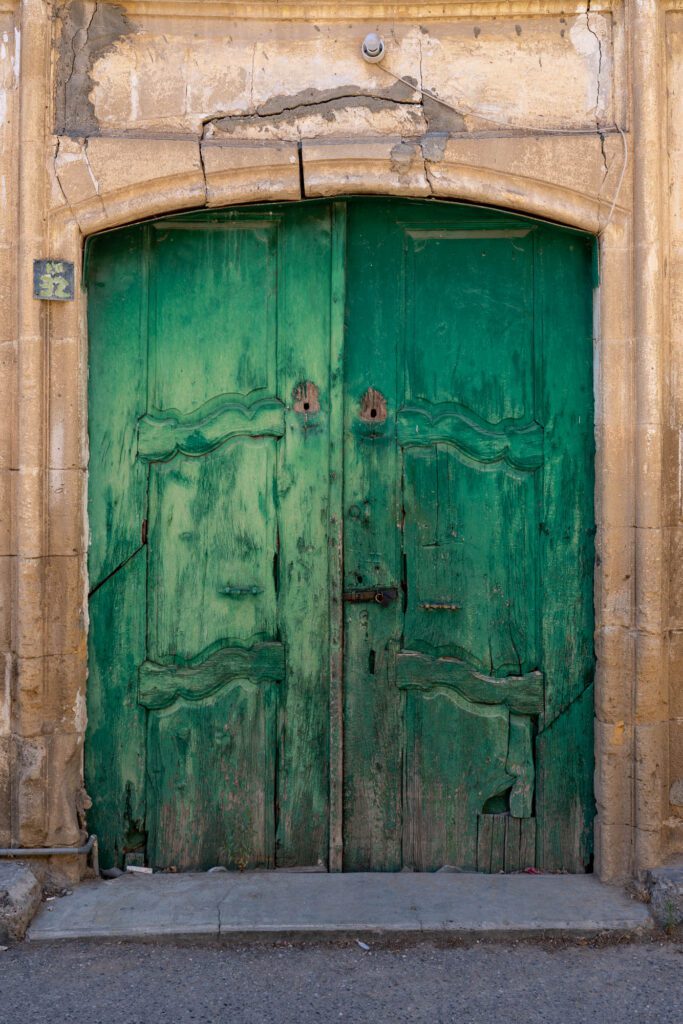 The image shows weathered green double doors set in a stone archway, exhibiting signs of age and erosion, with a rustic and antique appearance.