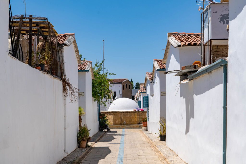 A narrow alley with white-walled buildings under a blue sky. Paved path leading to an archway. Rooftop tiles visible, possibly a Mediterranean setting.