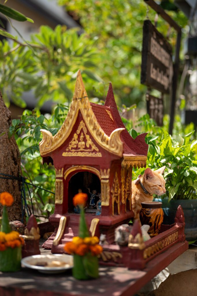 A decorated spirit house with ornate designs stands outdoors, surrounded by greenery. A cat rests nearby, creating a serene and picturesque scene.