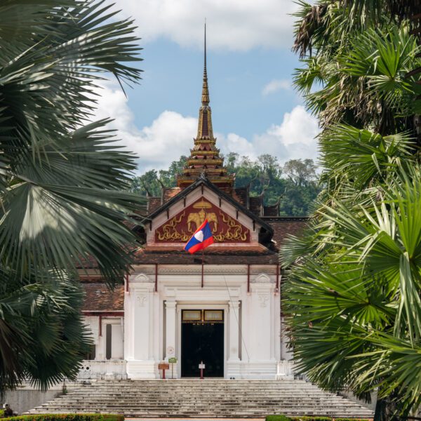 A path leads to the Royal Palace Museum in Luang Prabang, framed by palm trees under a blue sky with scattered clouds.